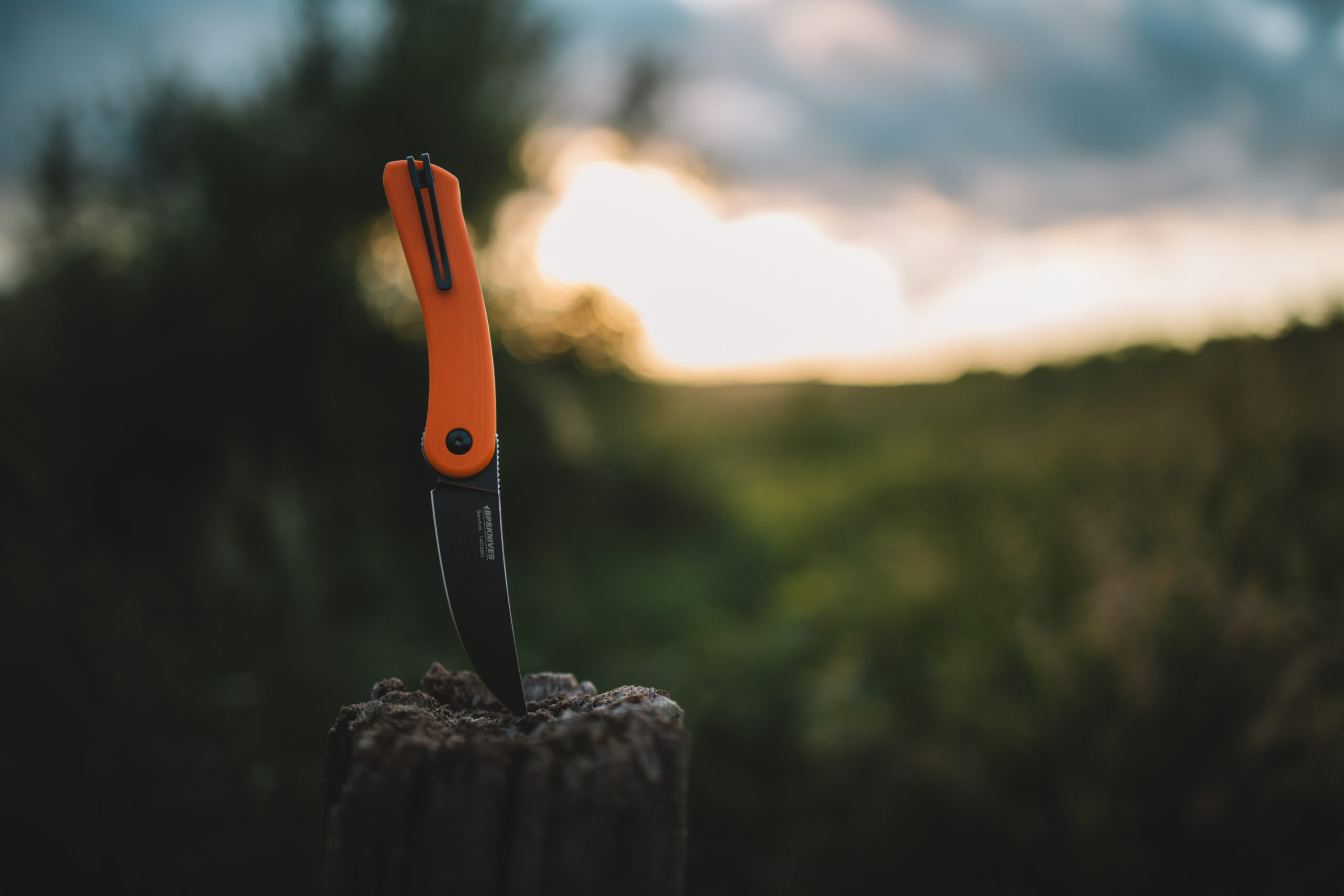 Folding knife with orange handle and black blade on a wooden post against a blurred natural background